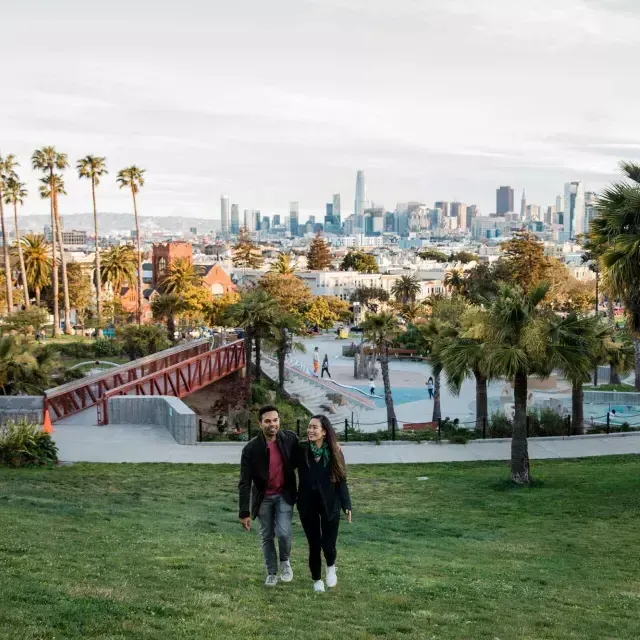 Ein Paar geht auf die Kamera zu, Dolores Park und die Skyline von San Francisco im Rücken.